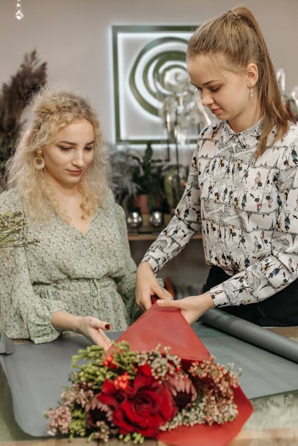 A florist from Ardleigh Green Florist wrapping a large, vibrant floral arrangement featuring deep red roses, delicate pink filler flowers, and green foliage with a red wrapping paper. The bouquet is p