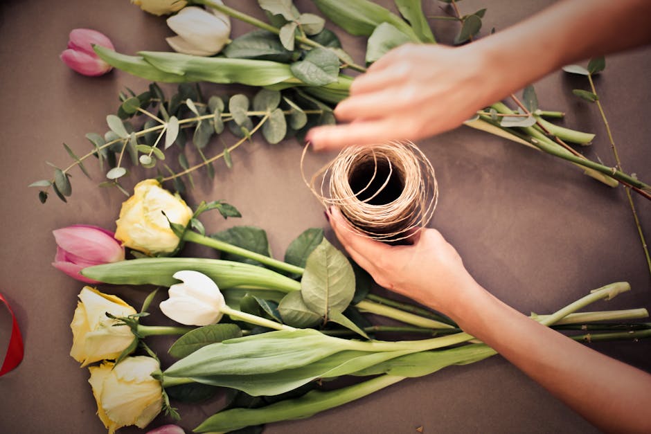 A florist is arranging a floral gift consisting of freshly cut roses in shades of yellow, white, and pink, along with eucalyptus and other greenery, on a workspace with a neutral surface. The florist'