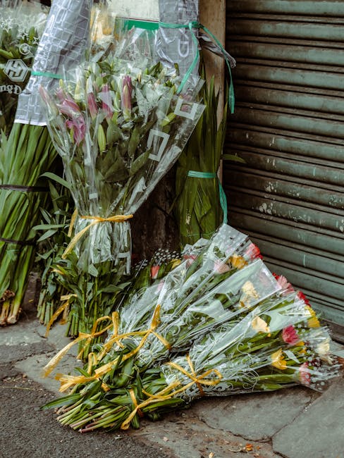 Multiple bouquets of fresh flowers, including pink lilies and yellow-tipped roses, wrapped in clear cellophane with decorative ribbons, are arranged on a pavement outside a building with a closed meta